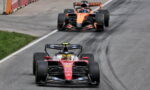 Two Formula 1 cars race on a track, red Ferrari leading followed by an orange McLaren with tire marks on the pavement behind them.