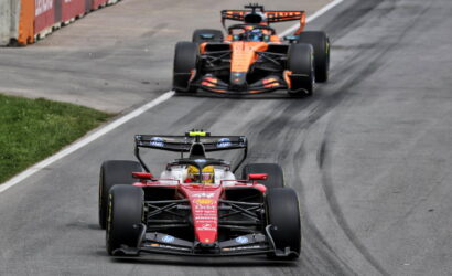 Two Formula 1 cars race on a track, red Ferrari leading followed by an orange McLaren with tire marks on the pavement behind them.
