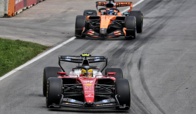 Two Formula 1 cars race on a track, red Ferrari leading followed by an orange McLaren with tire marks on the pavement behind them.