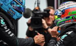 Two race car drivers in colorful helmets shake hands before a race, with a cameraman filming in the background.