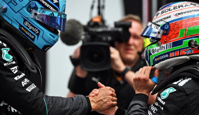 Two race car drivers in colorful helmets shake hands before a race, with a cameraman filming in the background.