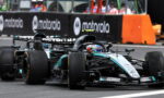 Two Mercedes Formula 1 cars race side by side on a track, with orange barriers and team personnel in the background.