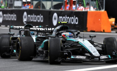 Two Mercedes Formula 1 cars race side by side on a track, with orange barriers and team personnel in the background.