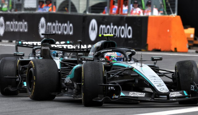 Two Mercedes Formula 1 cars race side by side on a track, with orange barriers and team personnel in the background.