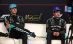 Two Mercedes Formula 1 drivers sit on a white couch in Montreal, wearing black racing suits with teal accents and blue caps.