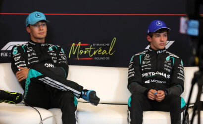Two Mercedes Formula 1 drivers sit on a white couch in Montreal, wearing black racing suits with teal accents and blue caps.