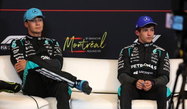 Two Mercedes Formula 1 drivers sit on a white couch in Montreal, wearing black racing suits with teal accents and blue caps.