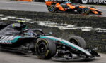 Mercedes–AMG Petronas Formula 1 car in foreground on a race track with an orange racing car in the background; sponsors visible include Microsoft, Petronas and IWC on the car body, with tire marks on the asphalt edge of the track.