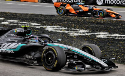 Mercedes–AMG Petronas Formula 1 car in foreground on a race track with an orange racing car in the background; sponsors visible include Microsoft, Petronas and IWC on the car body, with tire marks on the asphalt edge of the track.