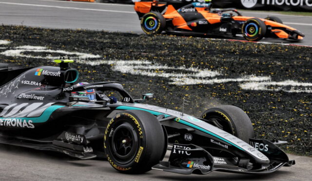 Mercedes–AMG Petronas Formula 1 car in foreground on a race track with an orange racing car in the background; sponsors visible include Microsoft, Petronas and IWC on the car body, with tire marks on the asphalt edge of the track.