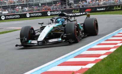 Mercedes F1 car (Petronas) charging through a sharp bend on a race track, turquoise and silver livery with a driver in blue helmet; stands and barriers in the background.