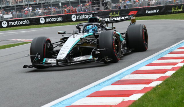 Mercedes F1 car (Petronas) charging through a sharp bend on a race track, turquoise and silver livery with a driver in blue helmet; stands and barriers in the background.