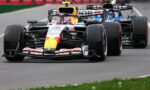 Two Formula One cars race closely on a track, the lead car in Red Bull livery with a yellow nose and sponsor logos