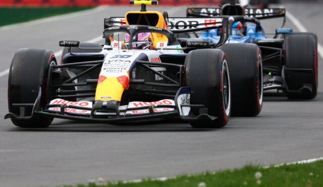Two Formula One cars race closely on a track, the lead car in Red Bull livery with a yellow nose and sponsor logos