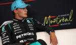 Male race car driver in a black Petronas suit and light blue cap sits on a white couch, Montreal backdrop behind him.