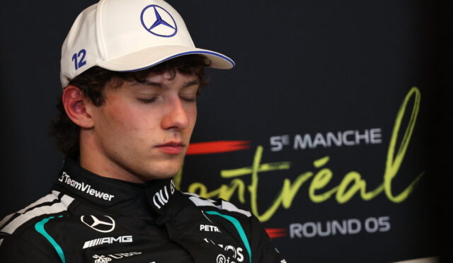 Young race car driver with eyes closed, wearing a white Mercedes cap and black racing suit with team logos, during a press event.
