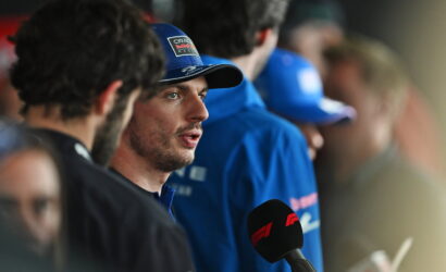 Driver wearing blue cap speaks to press, microphone with red F1 logo in foreground.