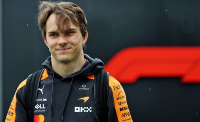 Smiling young man with a backpack wearing a black and orange team hoodie with logos, standing outdoors in front of a dark backdrop.