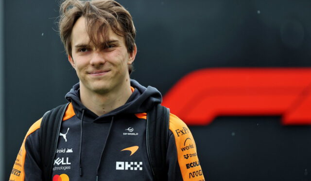 Smiling young man with a backpack wearing a black and orange team hoodie with logos, standing outdoors in front of a dark backdrop.