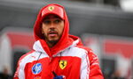 Man in a red and white Ferrari jacket and cap, looking to the side, holding a blue water bottle outdoors.
