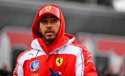 Man in a red and white Ferrari jacket and cap, looking to the side, holding a blue water bottle outdoors.