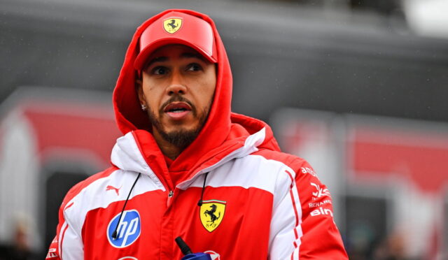 Man in a red and white Ferrari jacket and cap, looking to the side, holding a blue water bottle outdoors.