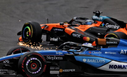 Two Formula 1 cars race side by side, orange and blue, with sparks flying from a tire as they speed along the track.