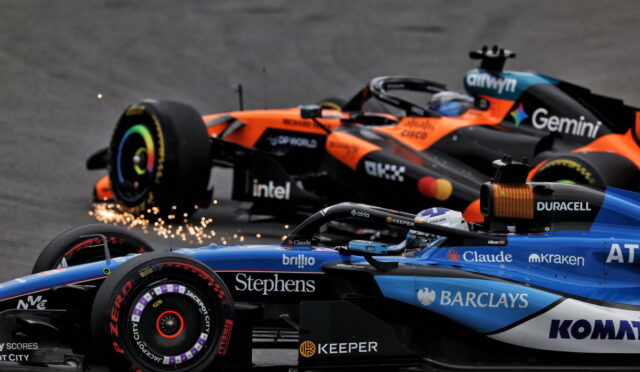 Two Formula 1 cars race side by side, orange and blue, with sparks flying from a tire as they speed along the track.