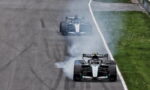 Two Mercedes Formula 1 cars drift on a race track, producing smoke as they power out of a corner on a grey tarmac with a grassy infield nearby.