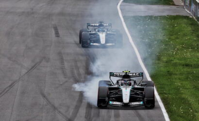 Two Mercedes Formula 1 cars drift on a race track, producing smoke as they power out of a corner on a grey tarmac with a grassy infield nearby.