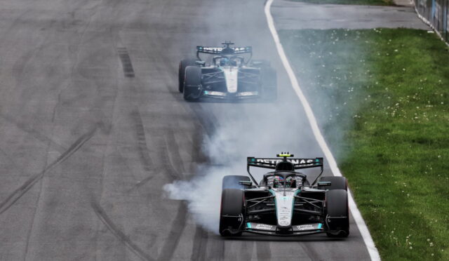 Two Mercedes Formula 1 cars drift on a race track, producing smoke as they power out of a corner on a grey tarmac with a grassy infield nearby.