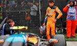 Driver in bright orange racing suit holds helmet beside a race car, with photographers and crew behind a fence in the background.