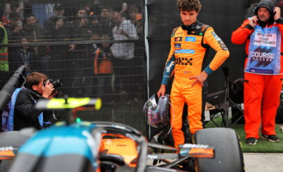 Driver in bright orange racing suit holds helmet beside a race car, with photographers and crew behind a fence in the background.