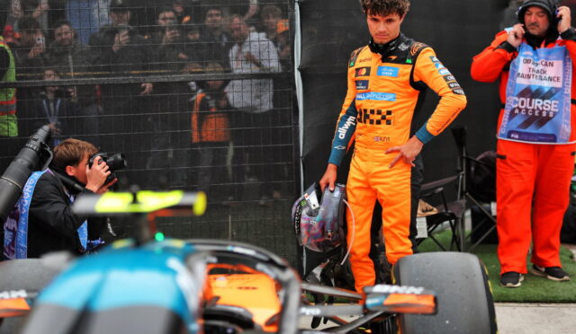 Driver in bright orange racing suit holds helmet beside a race car, with photographers and crew behind a fence in the background.