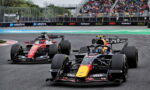 Front-running Red Bull Formula 1 car with a yellow nose leads a race, followed by a red Ferrari on the track, with a packed grandstand in the background.