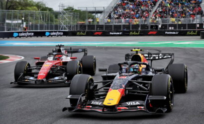 Front-running Red Bull Formula 1 car with a yellow nose leads a race, followed by a red Ferrari on the track, with a packed grandstand in the background.