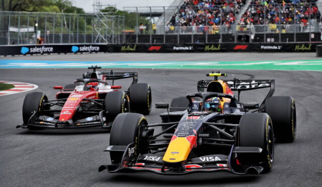 Front-running Red Bull Formula 1 car with a yellow nose leads a race, followed by a red Ferrari on the track, with a packed grandstand in the background.