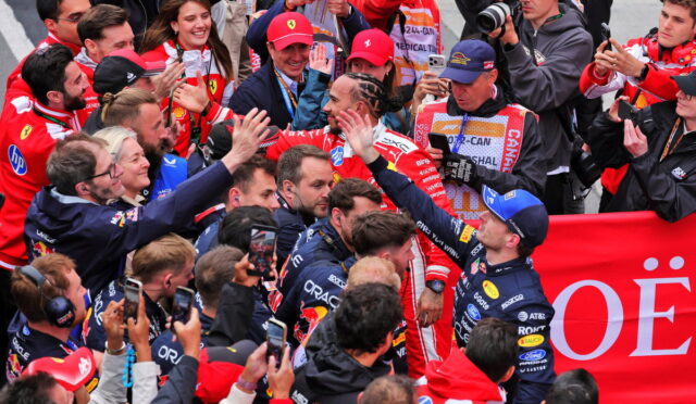 Group of Formula 1 drivers in racing suits exchanging high-fives amid cheering fans and photographers.