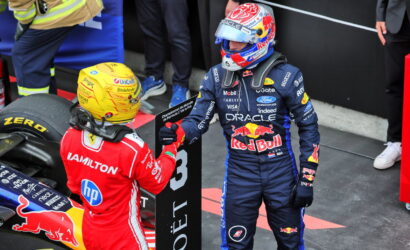 Two Formula 1 drivers in racing suits exchange a fist bump near a Red Bull car on the pit wall. The blue-clad driver stands, helmeted, beside the red-suited teammate and sponsor decals.