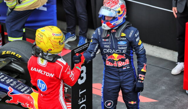Two Formula 1 drivers in racing suits exchange a fist bump near a Red Bull car on the pit wall. The blue-clad driver stands, helmeted, beside the red-suited teammate and sponsor decals.