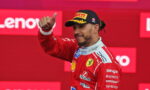 Ferrari Formula 1 driver on the podium giving a thumbs up, wearing a red racing suit and a Pirelli cap against a Lenovo backdrop.