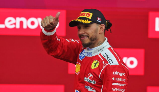 Ferrari Formula 1 driver on the podium giving a thumbs up, wearing a red racing suit and a Pirelli cap against a Lenovo backdrop.