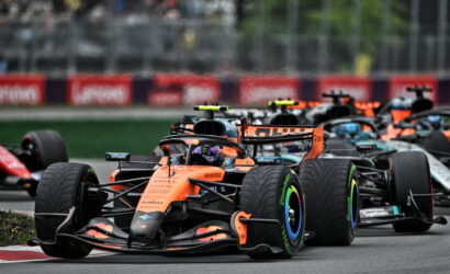 Orange Formula 1 car leading a pack on a race track with other cars close behind on the bend, blurred grandstands in the background.