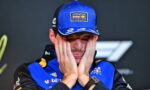 Race car driver in a blue Oracle Red Bull Racing cap and suit, hands pressed to his cheeks in a stressed expression.