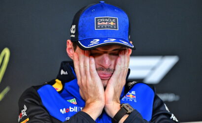 Race car driver in a blue Oracle Red Bull Racing cap and suit, hands pressed to his cheeks in a stressed expression.