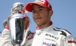 Race car driver in a white Renault racing suit and red cap posing with a large silver trophy against a blue sky.
