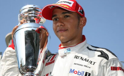Race car driver in a white Renault racing suit and red cap posing with a large silver trophy against a blue sky.