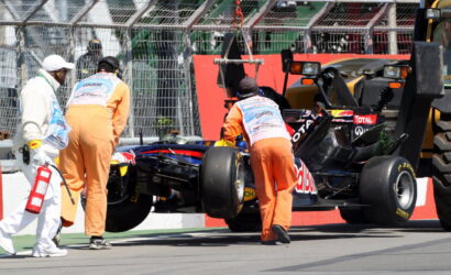 Pit crew in orange overalls push a Red Bull Formula 1 car while a safety worker in white carries a fire extinguisher nearby after an incident.