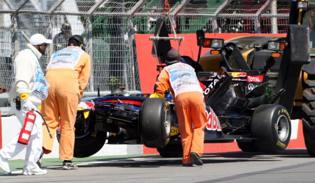 Pit crew in orange overalls push a Red Bull Formula 1 car while a safety worker in white carries a fire extinguisher nearby after an incident.