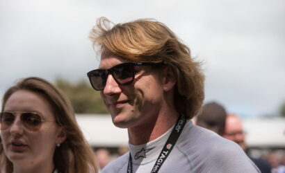 Man with light brown hair wearing dark sunglasses and a lanyard at an outdoor event; smiling slightly, with a woman wearing sunglasses beside him in the foreground.